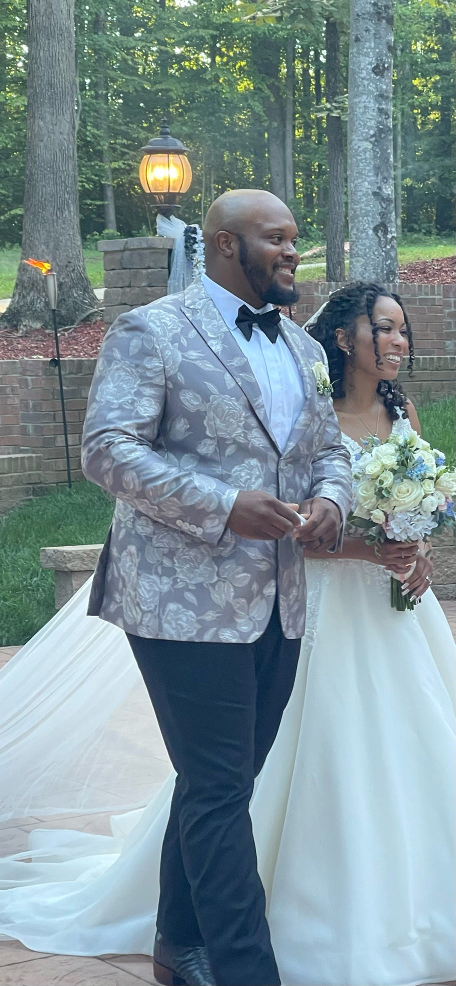 Groom in patterned suit and bride in white dress holding bouquet outdoors.